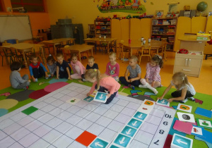 Grupa dzieci siedzi wokół maty ułożonej na dywanie i obserwuje jak dziewczynka w różowej bluzce układa trasę auta do zielonej tabliczki, obok na macie ułożona jest już jedna trasa do czerwonej tabliczki.